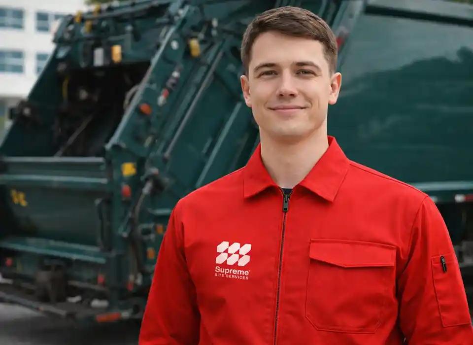 A man in a red uniform stands smiling in front of a garbage truck.